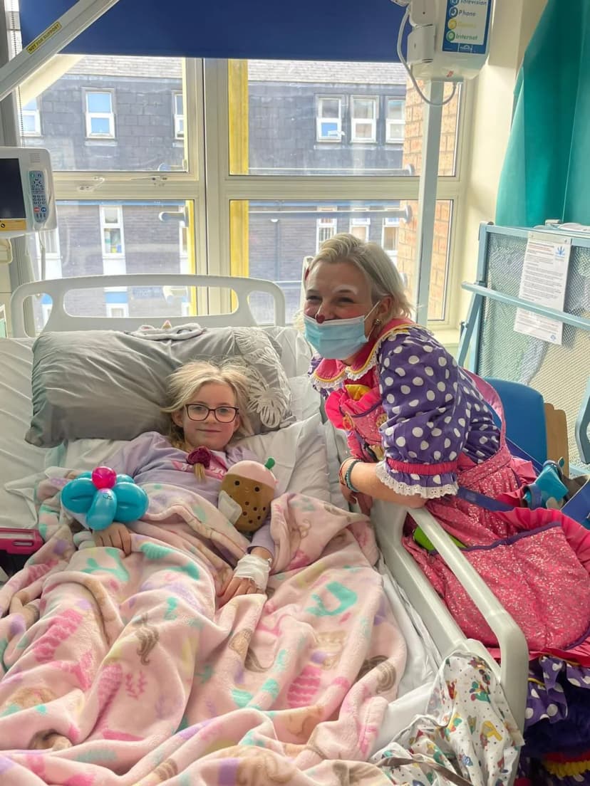 This is an image of Louby Lou at a hospital smiling for a photo with a child patient in her clown attire.