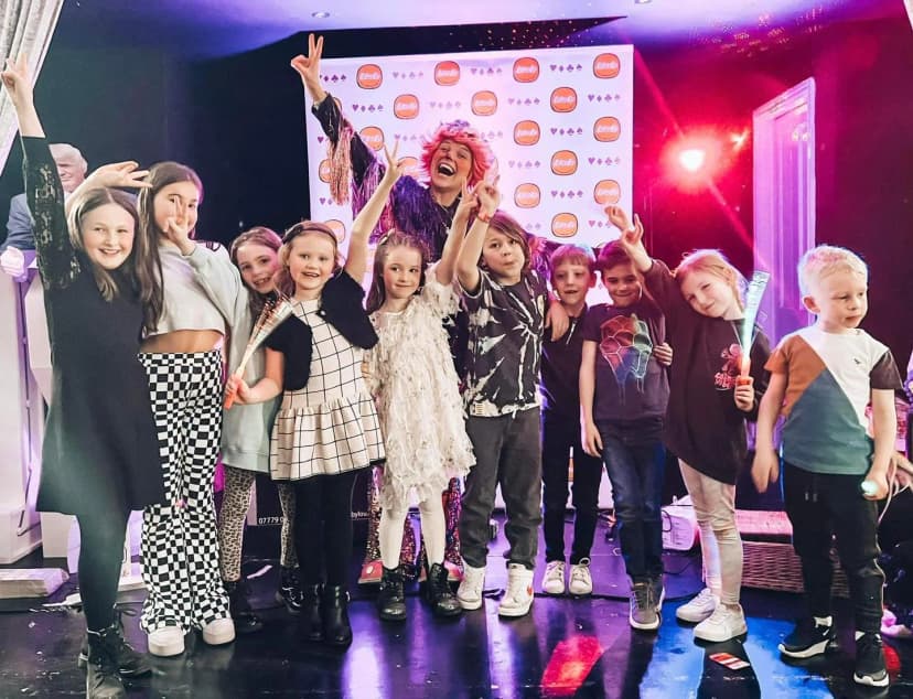An image of Louby Lou, the clown, posing in front of a Louby Lou banner with a group of 10 children at a party. Each child is giggling and celebrating. The image really radiates excitment. This image was taken in Greater Manchester